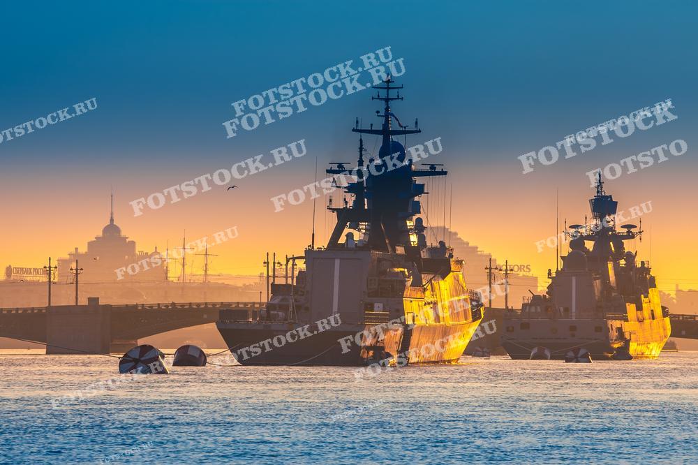 На фотографии изображены военные корабли, стоящие на реке Нева в Санкт-Петербурге, на фоне восхода солнца. Свет утреннего солнца отражается на воде и на корпусах кораблей, создавая красивое золотистое свечение. На заднем плане виднеется мост и городские постройки, скрытые в утреннем тумане.